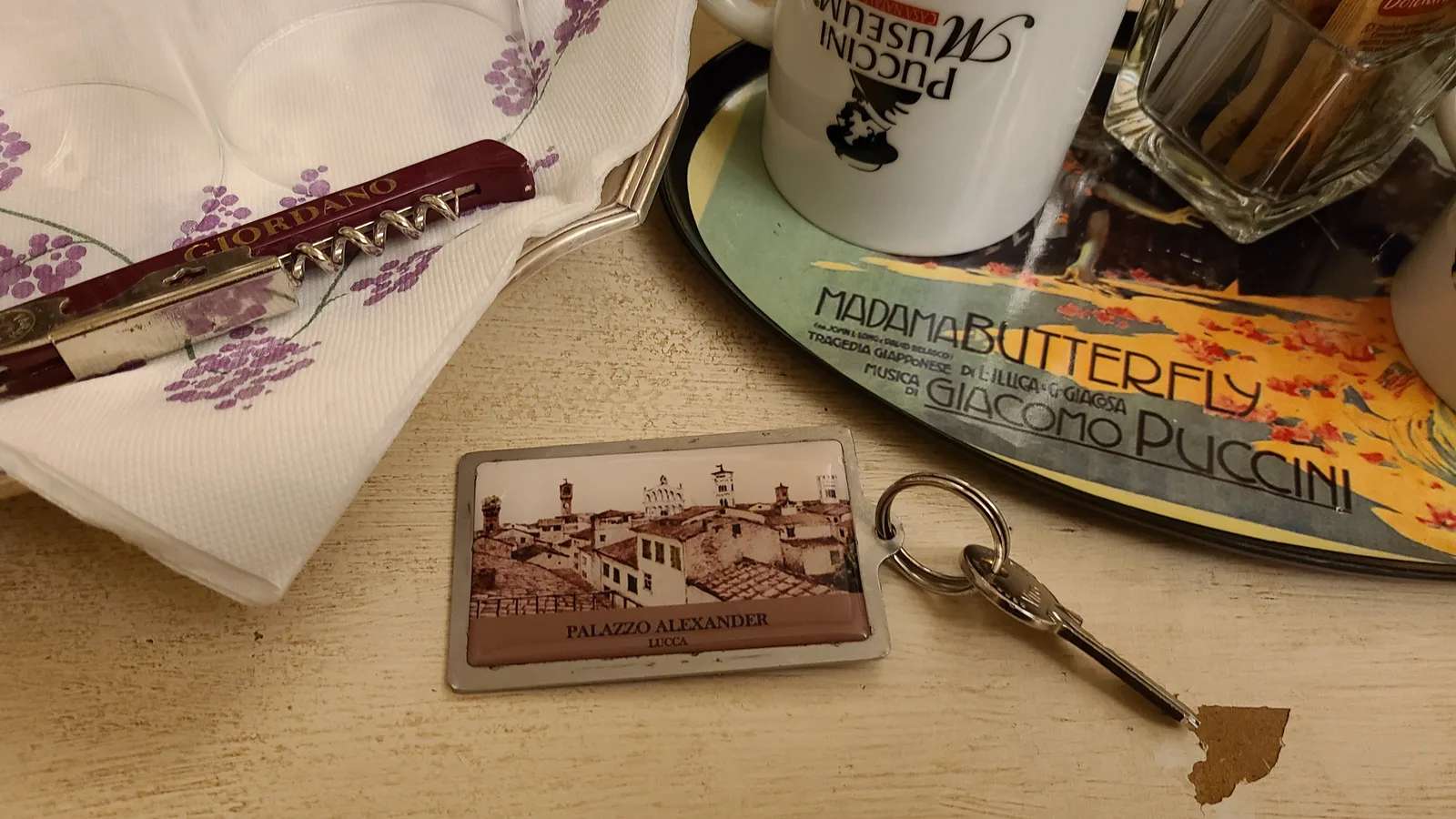 Hotel Palazzo Alexander room key with a Lucca cityscape engraving on the keychain, resting next to a Puccini museum coffee cup and Madama Butterfly program