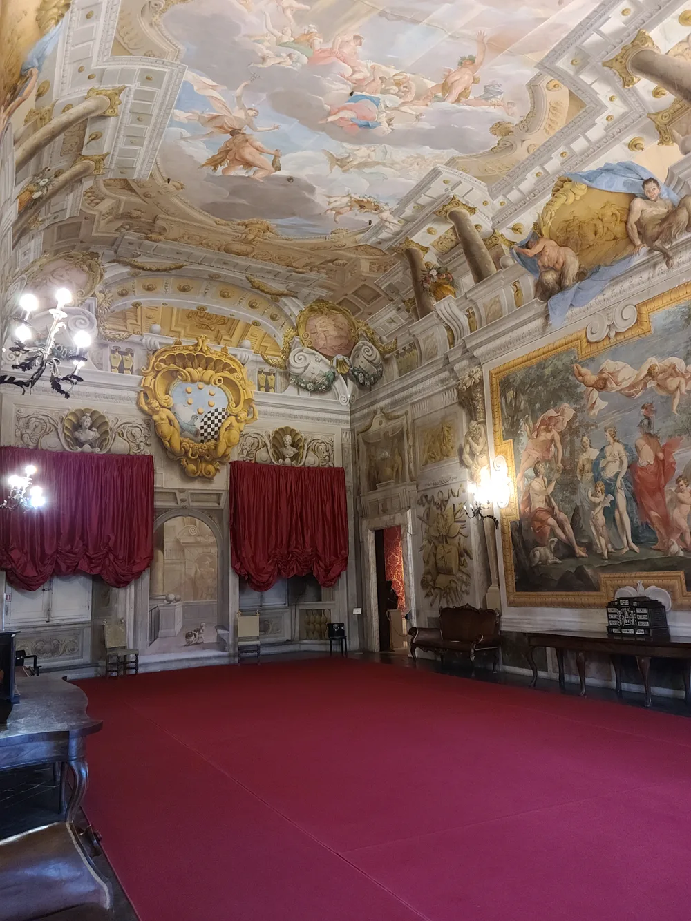 Baroque state room in a Lucca palazzo with an elaborate ceiling fresco of mythological figures in flight, red velvet drapes, and a large oil painting on the wall