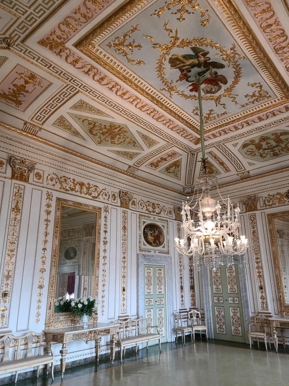 Opulent gilded ballroom inside a Lucca palazzo with an intricate gold-and-white decorated ceiling, crystal chandelier, large gilt mirror, and period chairs along the walls