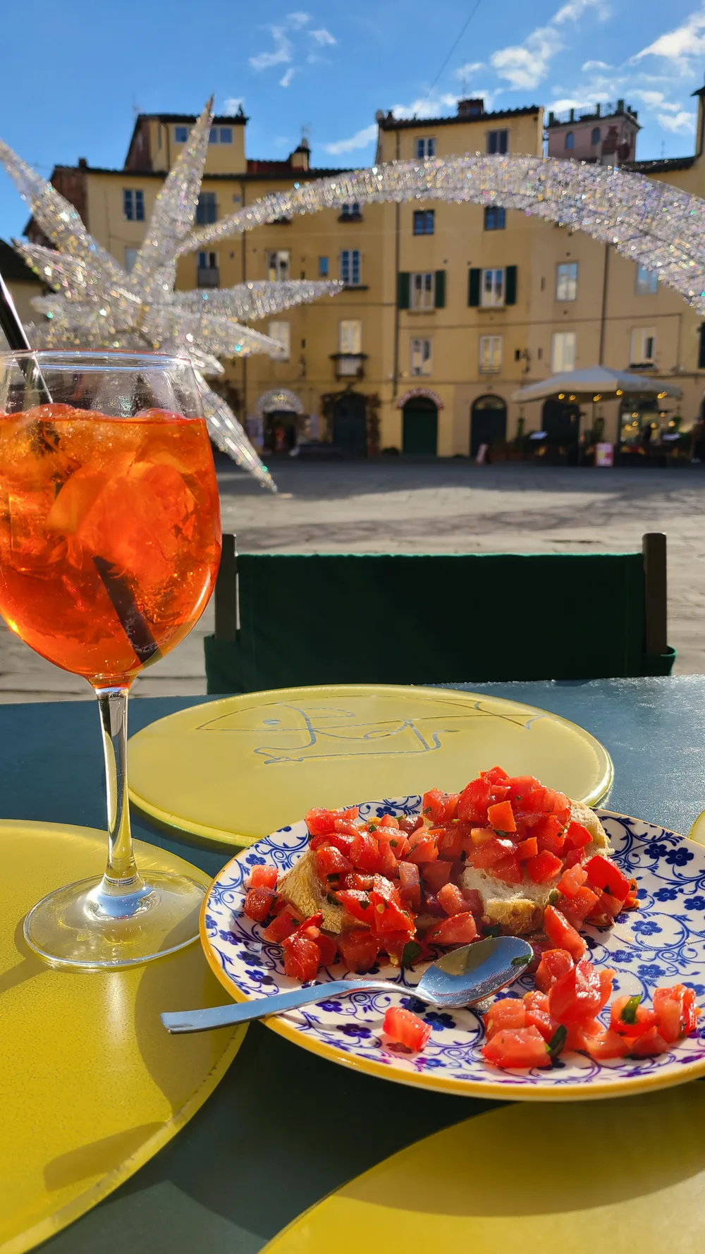 Aperol Spritz and bruschetta on a yellow café table at Piazza dell'Anfiteatro, Lucca, colorful medieval buildings and Christmas decorations in the background