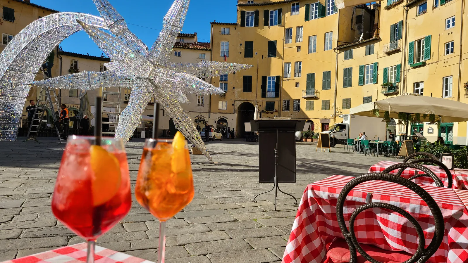 Two Aperol Spritz drinks on a café table at Piazza dell'Anfiteatro, Lucca, with glittering Christmas star decorations and the oval ring of medieval buildings behind
