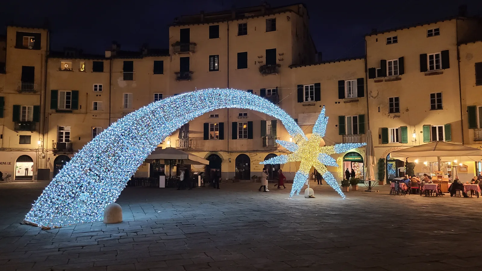 Piazza dell'Anfiteatro in Lucca at night with a large illuminated shooting-star Christmas light sculpture in the oval medieval square, surrounding buildings warmly lit