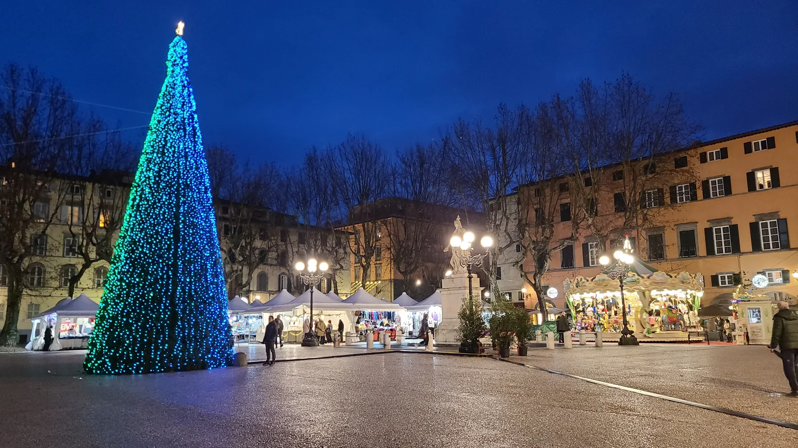 Piazza Napoleone in Lucca at dusk with a tall illuminated Christmas tree, market stalls and a carousel in front of the surrounding buildings lit in the evening