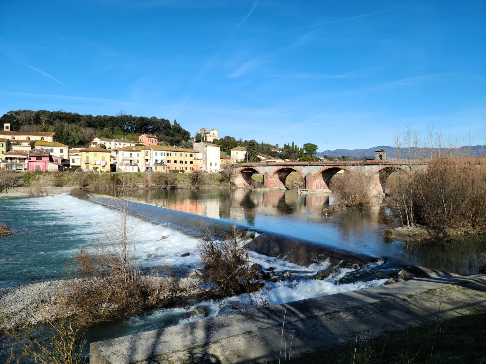 Multi-arched stone bridge at Ponte a Moriano spanning the fast-flowing Serchio River, a colorful village on a wooded hillside beyond under a blue sky