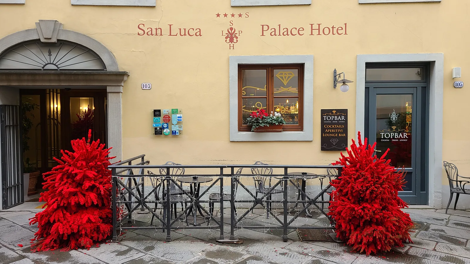 San Luca Palace Hotel entrance in Lucca with two bold red feather Christmas tree sculptures flanking the doorway, a TopBar sign visible alongside