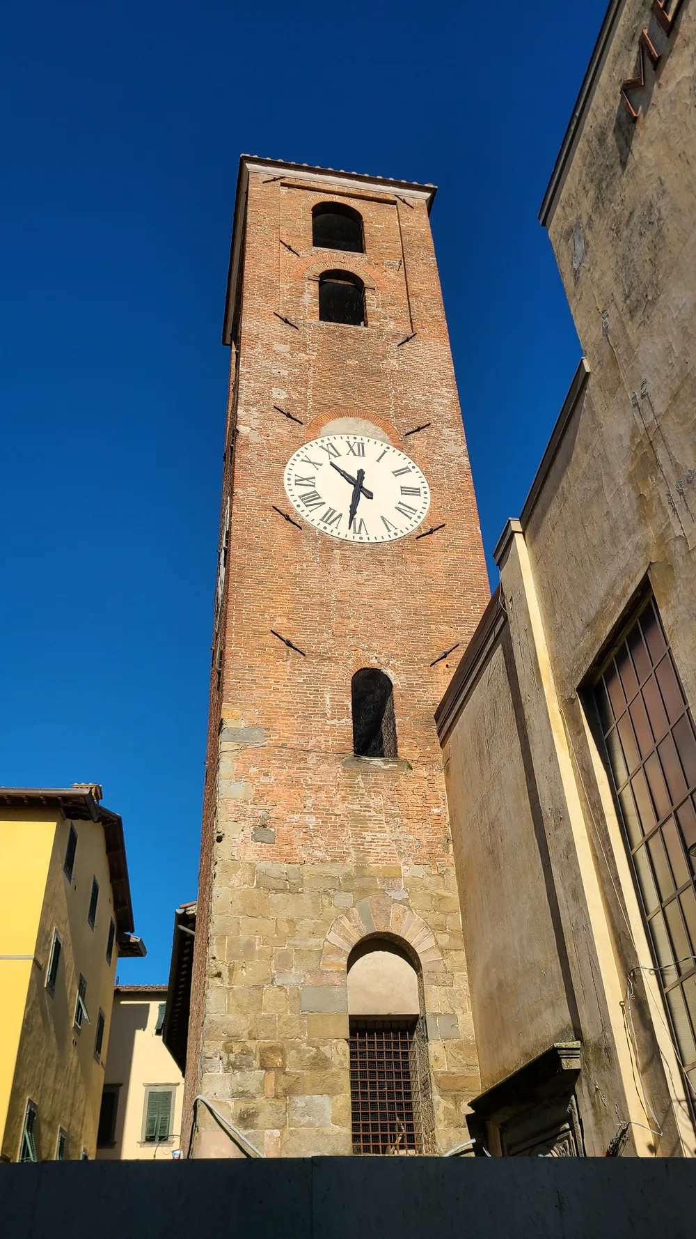 Torre delle Ore medieval brick clock tower in Lucca seen from street level, the white clock face and tower rising against a deep blue sky