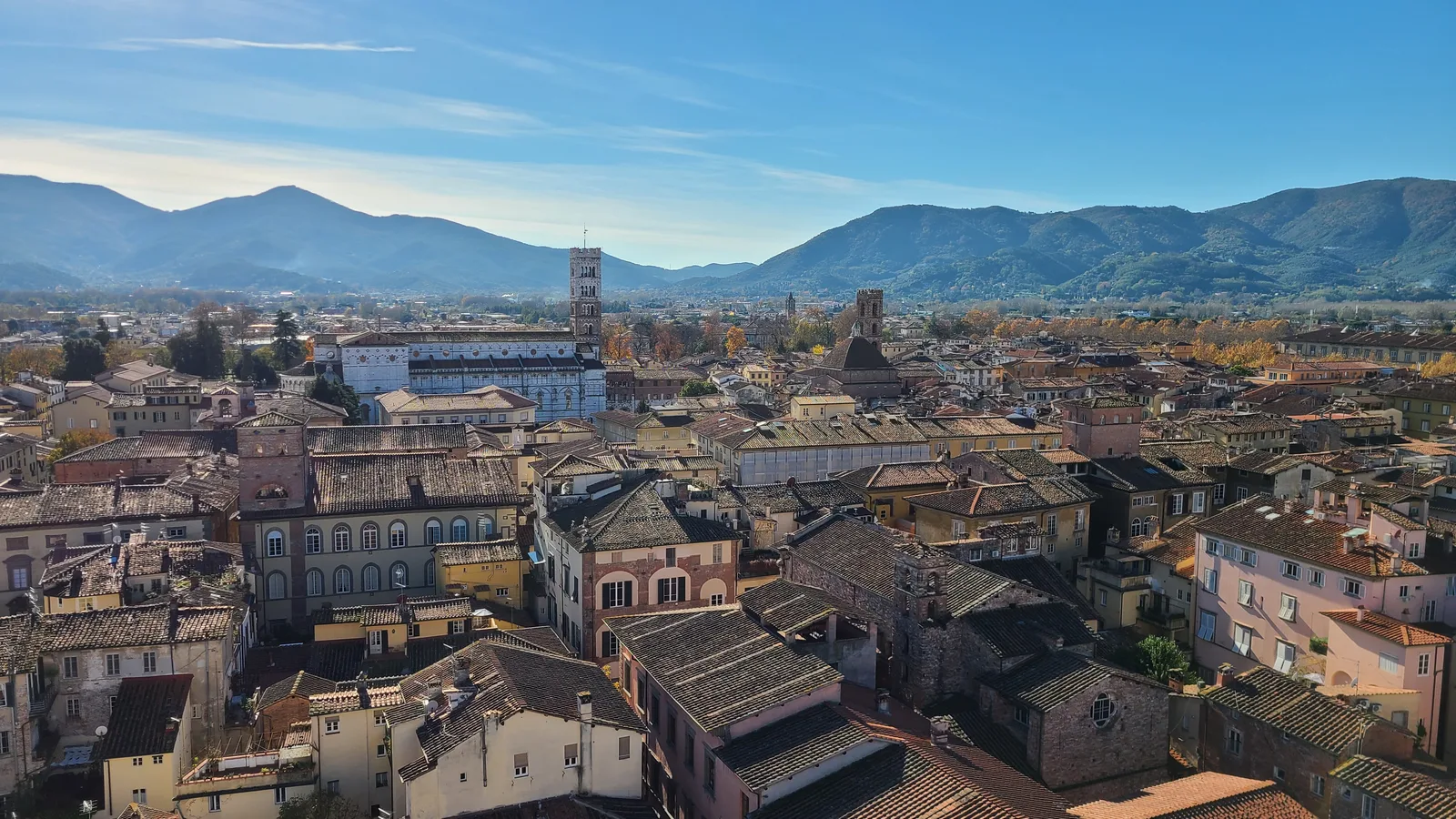 Panoramic view from the top of Torre Guinigi in Lucca, terracotta rooftops spreading across the city with Torre delle Ore and the Apuan Alps visible in the distance