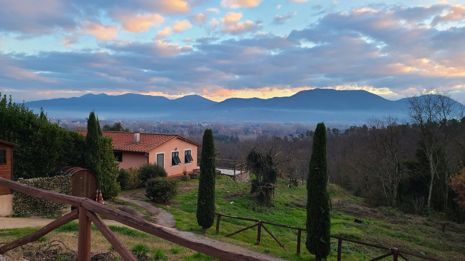 Tuscan countryside near Lucca at dusk, cypress trees and an orange-roofed farmhouse in the foreground with the Apuane Alps silhouetted against a dramatic sunset sky
