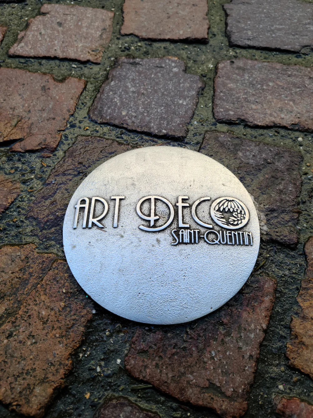 Art Deco bronze heritage marker embedded in cobblestones on a Saint-Quentin pedestrian street