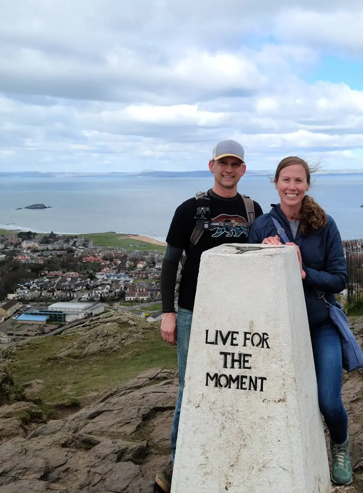 Andrew and Ashley at the summit of North Berwick Law beside the trig point reading 'Live For The Moment', with the town of North Berwick and the Firth of Forth stretching out below